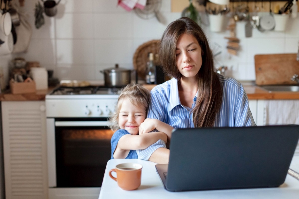Woman at kitchen table with laptop and smiling child hugging her arm
